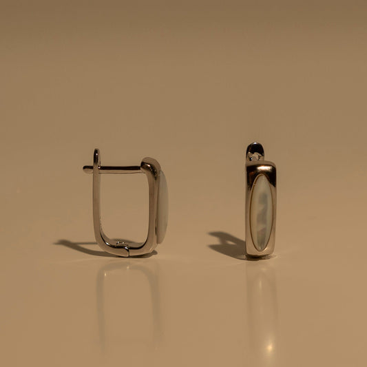 A pair of silver-toned metal hoop earrings with a smooth, polished finish, displayed on a reflective beige surface against a matching beige background.