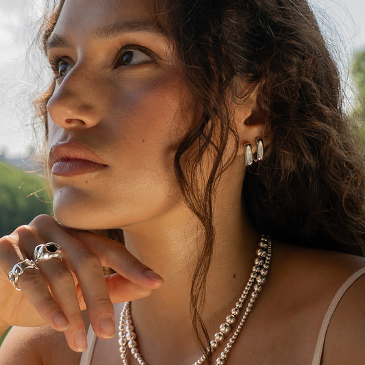 A close-up lifestyle shot of a woman with dark curly hair wearing silver jewelry, including a double-strand pearl necklace, a silver hoop earring, and silver rings, set against a blurred background of green trees and city buildings.
