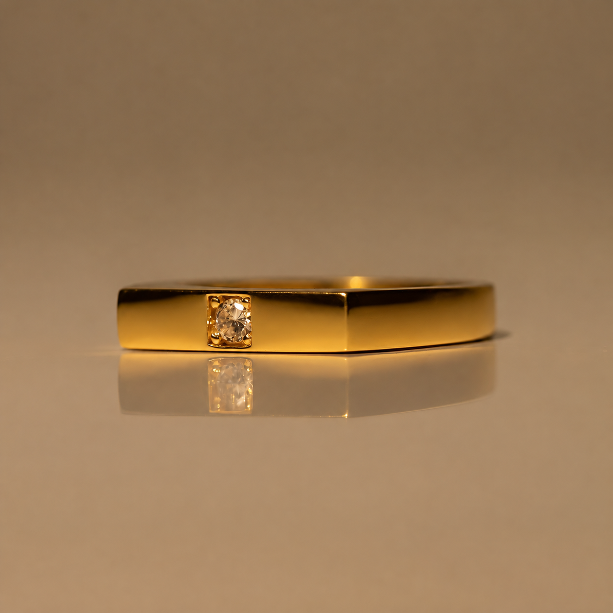 Front view of the Milk & Rose Agathe Adjustable Ring, a polished gold-tone rectangular band set with a single small clear gem, on a reflective beige surface.