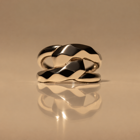Front view of the Milk & Rose Bold Unity Ring, a polished gold-tone double band with two overlapping faceted wave-shaped sections, on a reflective beige surface.