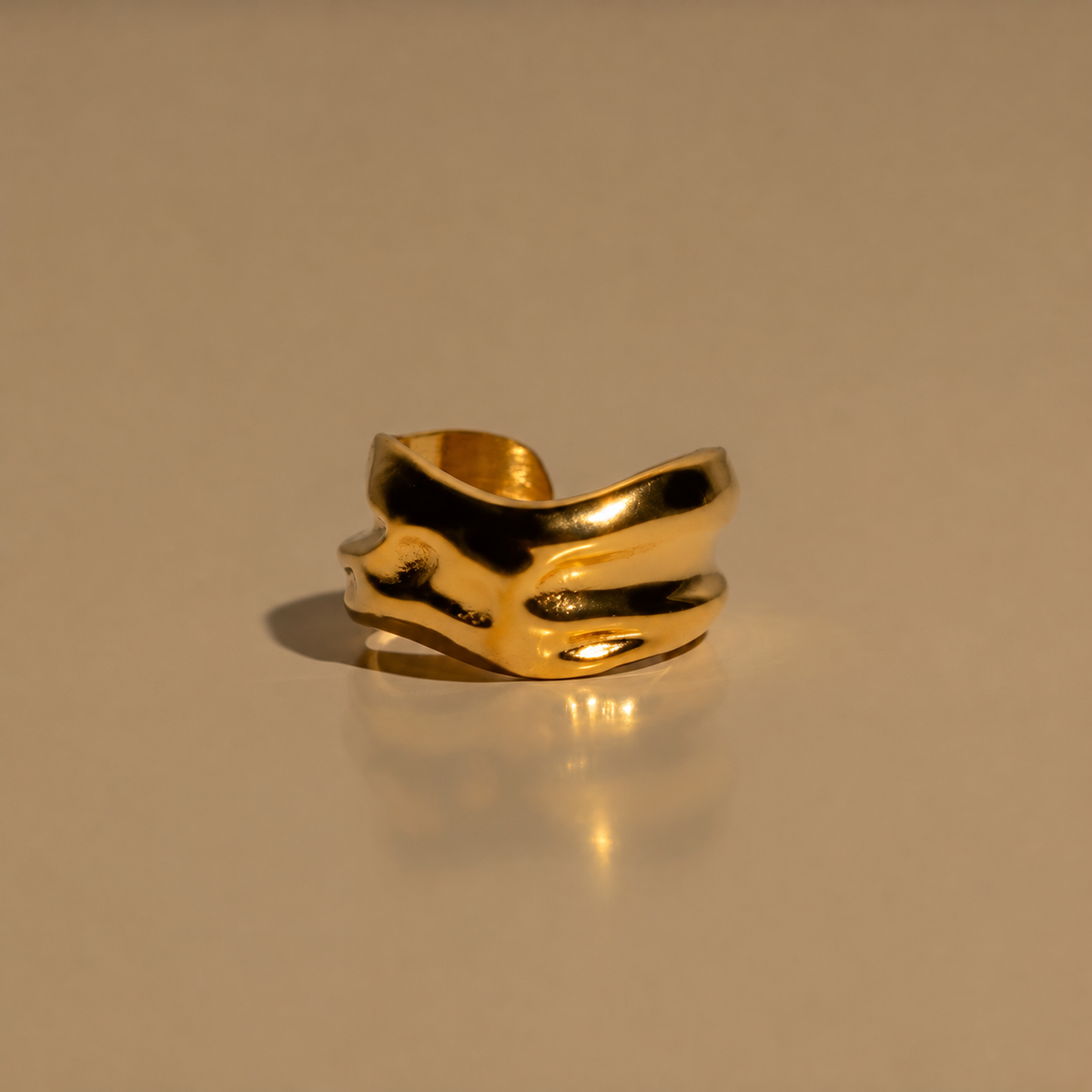 Front view of the Milk & Rose Circe Cuff on a reflective beige surface, showing a broad gold-tone band with flowing raised curves and an open back.