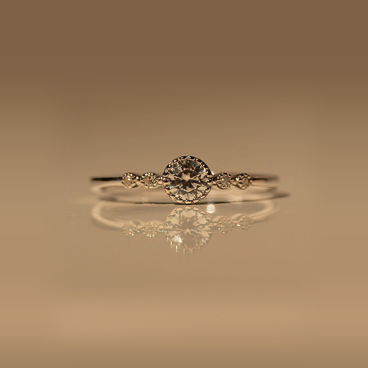 Front view of the Milk & Rose Bubble Gem Ring, a slim silver-tone band with a round center gem in a beaded setting and small accent stones on each side, on a reflective beige surface.