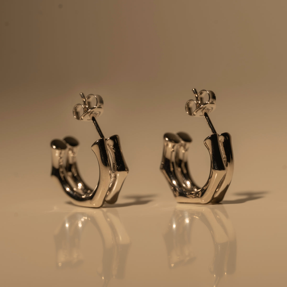 Milk & Rose Eve Hoops in a silver tone, with flat angular curves and butterfly backs, standing together on a reflective surface.