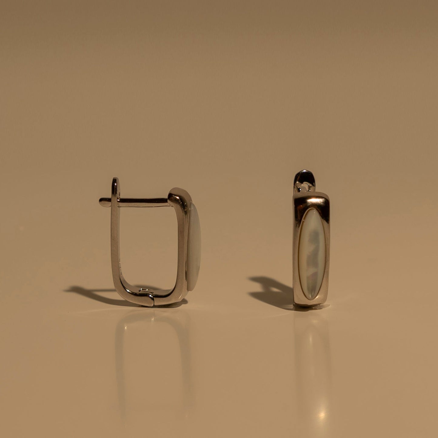 A pair of silver-toned metal hoop earrings with a smooth, polished finish, displayed on a reflective beige surface against a matching beige background.