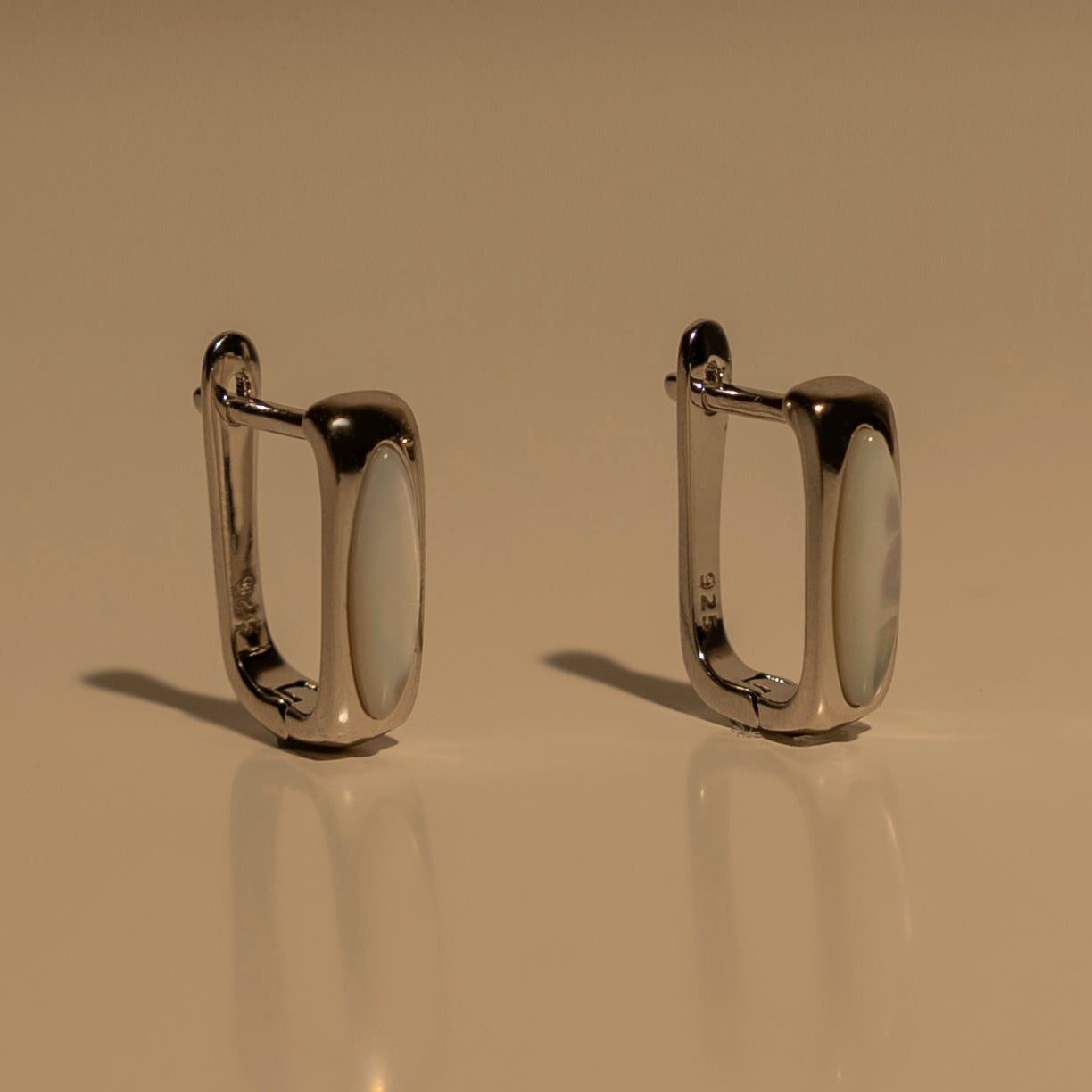 A pair of polished moonstone and silver-toned metal hoop earrings with a rounded rectangular design, displayed on a reflective beige surface.